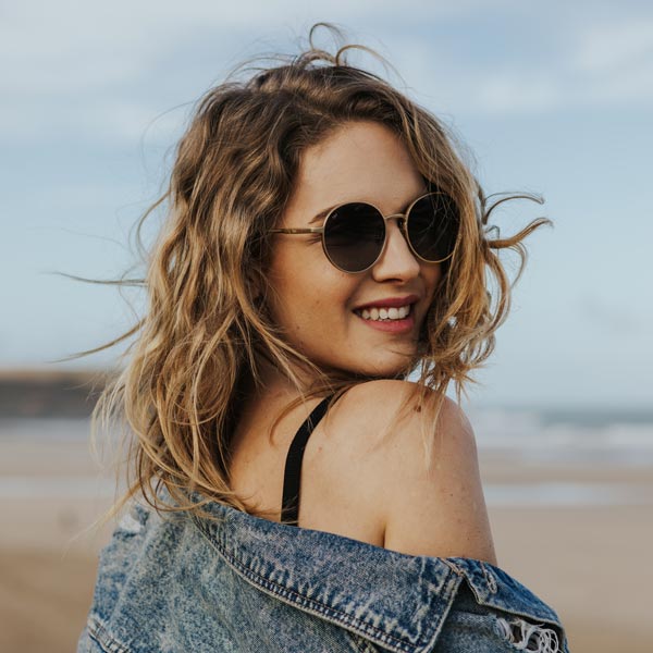 Blonde girl on a beach wearing  round sunglasses with gold frame and grey lenses