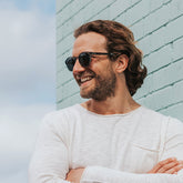 Man smiling standing against wall wearing clear grey frame sunglasses with polarised lenses